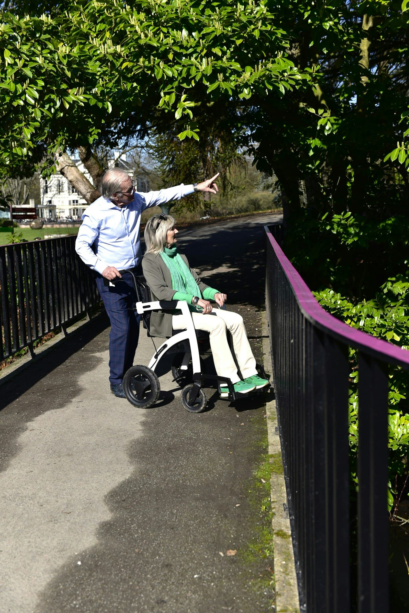 An older couple out on a sunlit tarmac path with handrails, one walking and pushing the other in a wheelchair, both looking up at something he is pointing to.