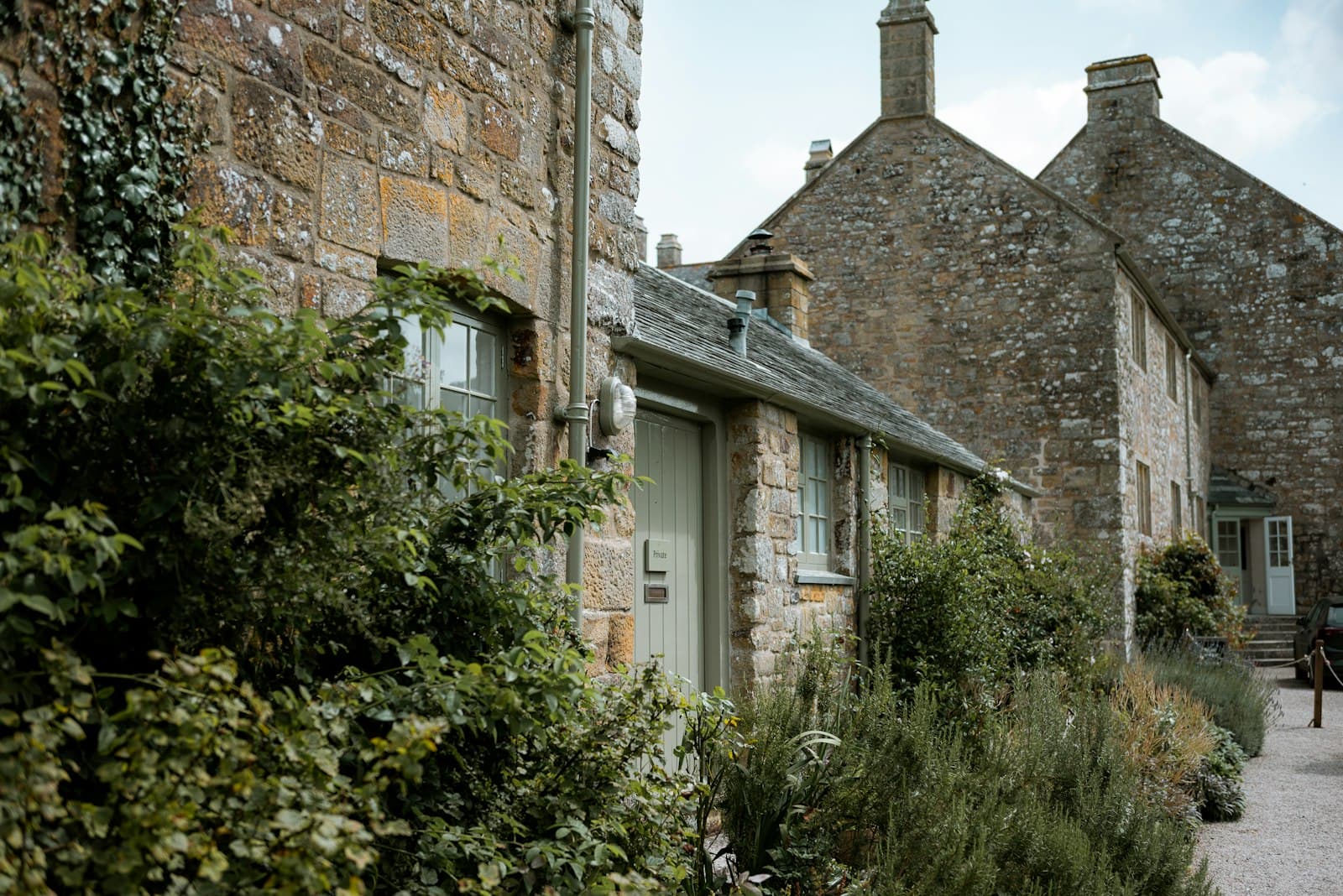 A row of stone cottages with a sage-green front door, beds of herbs and shrubs, and a gravel path running alongside.